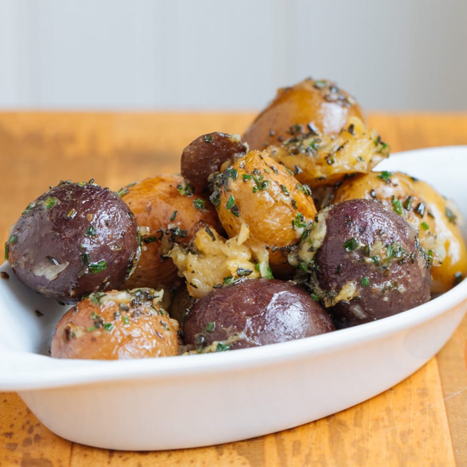 A white bowl filled with roasted brown and purple potatoes, garnished with herbs and garlic.