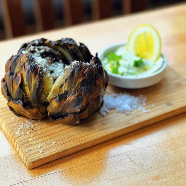 Grilled artichoke with parmesan cheese, a side of sauce, and lemon on a wooden board.