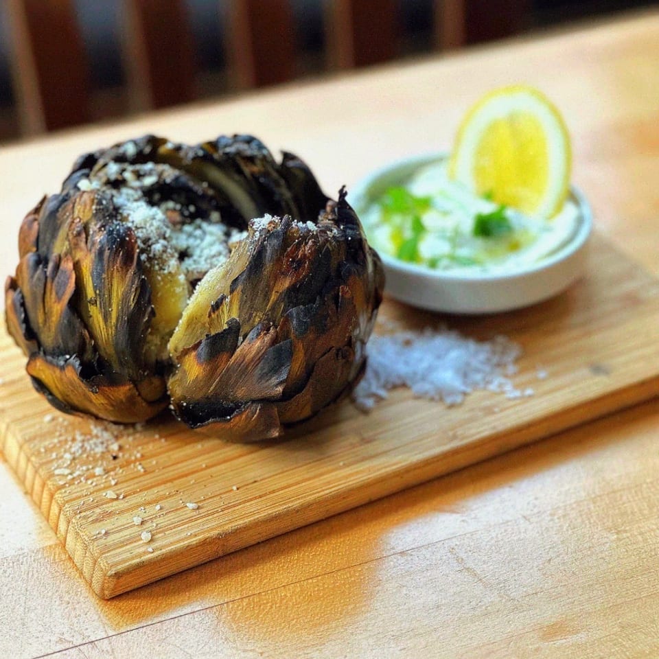 Grilled artichoke with parmesan cheese, a side of sauce, and lemon on a wooden board.