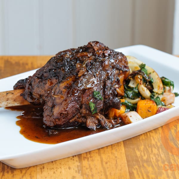 Braised lamb shank with rich brown sauce, served with roasted vegetables on a white plate.