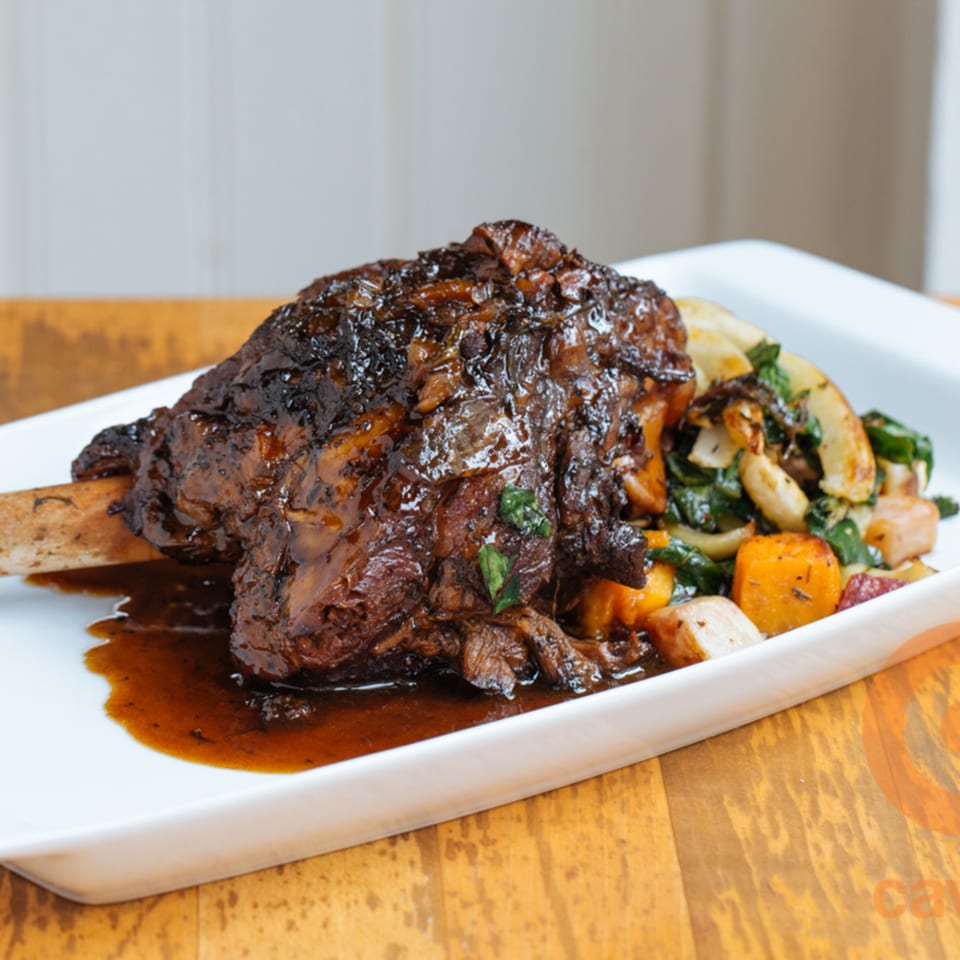Braised lamb shank with rich brown sauce, served with roasted vegetables on a white plate.