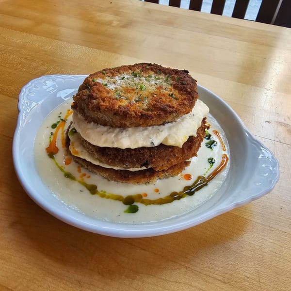 Stacked fried eggplant parmesan with creamy sauce and herb oil on a white plate.